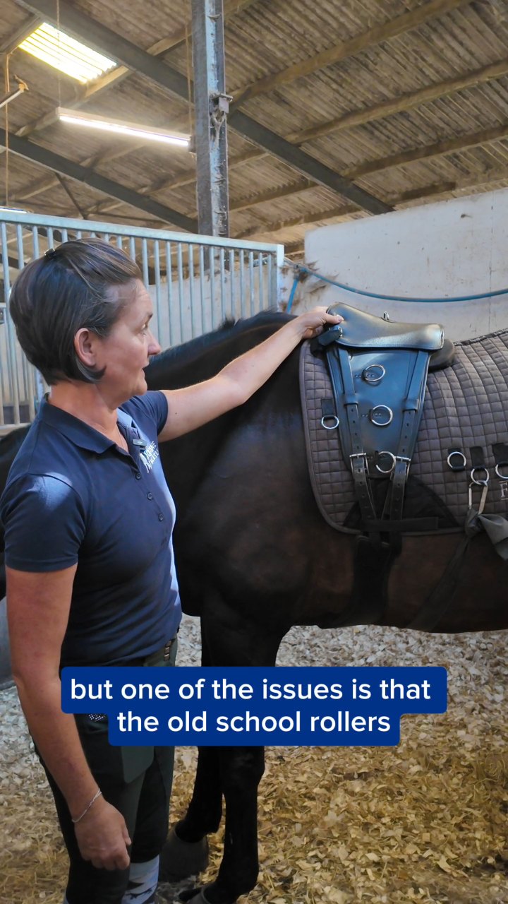 A dark horse in a barn wears a quilted pad and leather roller. A woman in a navy polo stands at its shoulder, talking to camera.